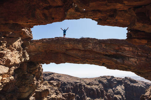 Junger Mann mit ausgestreckten Armen, der auf einem natürlichen Felsbogen steht, Gran Canaria, Spanien - RSGF00749