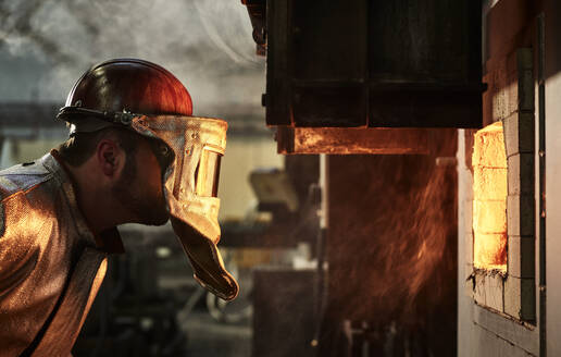 Arbeiter mit Schutzhelm schaut auf einen brennenden Ofen in der Industrie - CVF01768