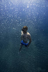Hemdloser Mann mit Schwimmbrille schwimmt inmitten von Blasen unter Wasser - RSGF00760