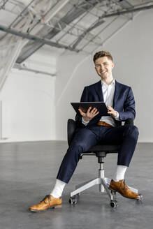 Lächelnder Geschäftsmann mit Tablet-PC auf einem Stuhl in einer Industriehalle - PESF03395