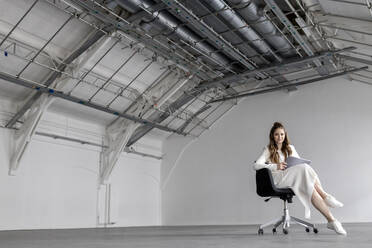 Geschäftsfrau sitzt mit Tablet-PC auf einem Stuhl in einer Fabrik - PESF03401