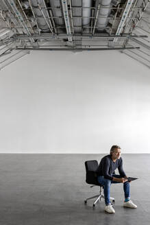 Geschäftsmann mit Tablet-PC auf einem Stuhl in einer leeren Industriehalle sitzend - PESF03415