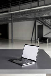 Laptop mit leerem Bildschirm auf einem Schreibtisch in einer Industriehalle - PESF03433