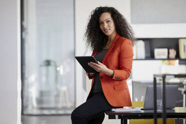 Lächelnde Geschäftsfrau, die einen Tablet-PC hält und auf einem Schreibtisch im Büro sitzt - RBF08464