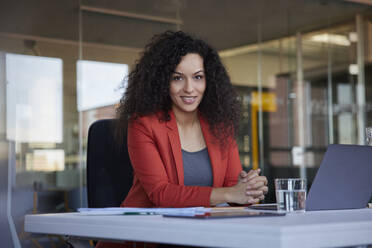 Smiling businesswoman with black curly hair at desk in office - RBF08480