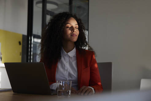 Businesswoman sitting with eyes closed in sunlight at workplace - RBF08493