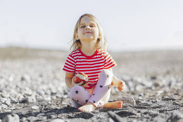 Mädchen hält Apfel am Strand sitzend an einem sonnigen Tag - OMIF00251