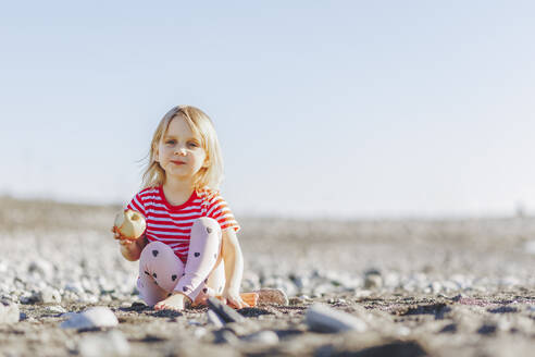 Nettes blondes Mädchen mit Apfel am Strand sitzend an einem sonnigen Tag - OMIF00252