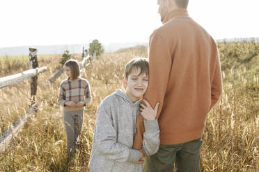 Smiling boy with eyes closed holding hand of father with sister in background - EYAF01831