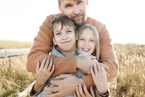 Vater umarmt lächelnd Tochter und Sohn an einem sonnigen Tag - EYAF01833