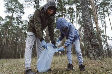 Father and son collecting plastic waste in garbage bag at forest - EYAF01837