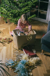 Girl searching ornaments in cardboard box sitting by Christmas tree at home - KMKF01780