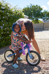 Mother kissing cute toddler daughter on bicycle in sunny driveway - CAIF32024