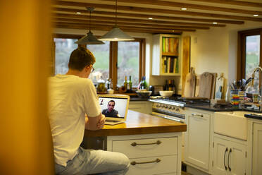 Man video conferencing with colleagues at laptop in kitchen - CAIF32075