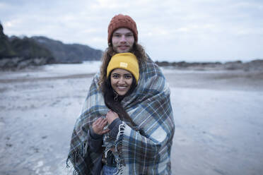 Portrait glückliches junges Paar in Decke eingewickelt am kalten Strand - CAIF32218