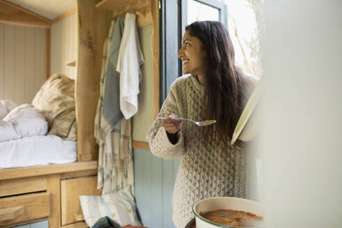 Happy young woman cooking stew in tiny cabin rental - CAIF32250