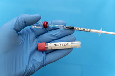 Doctor with protective glove holding swab tube with Omicron and syringe against blue background - DRF01783