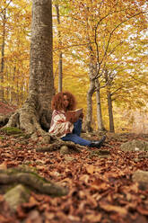 Frau, die ein Buch liest, sitzt an einem Baum im Herbstwald - VEGF05251