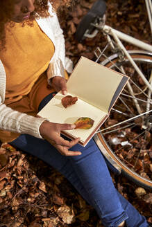 Frau hält Blatt in Buch sitzend am Fahrrad im Herbstwald - VEGF05274