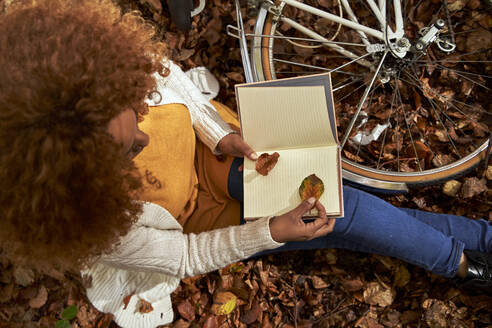 Frau mit Fahrrad hält Blatt in Buch sitzend im Herbstwald - VEGF05275