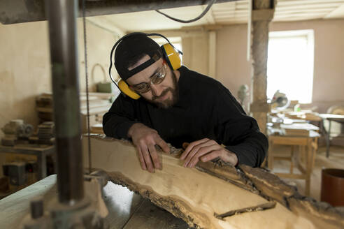 Handwerker mit Ohrenschützer beim Schnitzen von Holz durch eine Bandsäge in der Werkstatt - LLUF00440