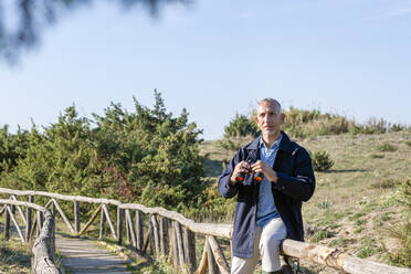 Man with binoculars sitting on railing at wooden bridge - EIF02799