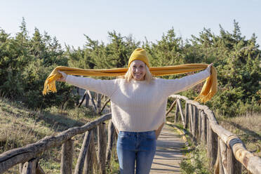 Smiling woman with arms outstretched holding scarf on wooden bridge - EIF02807