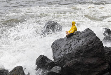 Young man wearing raincoat sitting on rock by waves splashing in sea, Rocha Da Relva, San Miguel Island, Azores, Portugal - WWF05937