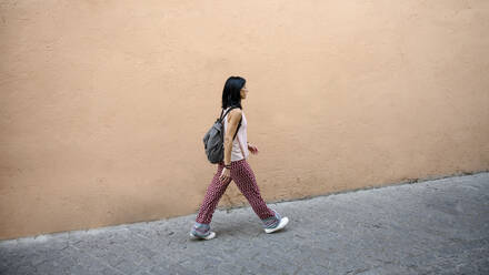 Woman with backpack waking on footpath by peach wall - RFTF00151