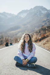 Carefree woman sitting cross-legged on road in front of mountains - OMIF00262