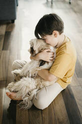 Boy embracing dog sitting on floor at home - SEAF00290