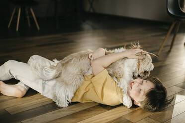 Smiling boy embracing dog and lying on hardwood floor at home - SEAF00292