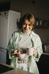 Lächelnde Frau mit Kaffeetasse und Schüssel in der Küche zu Hause - SEAF00293