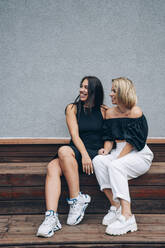 Smiling women sitting together on bench in front of wall - OMIF00267