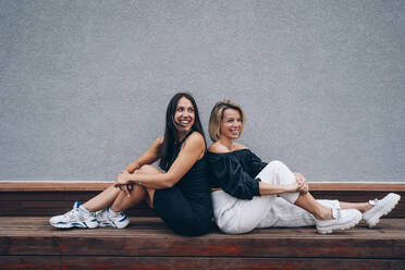Lächelnde Freunde sitzen Rücken an Rücken auf einer Bank vor einer Mauer - OMIF00272