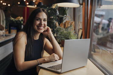 Junge Frau sitzt lächelnd mit Laptop in einem Café - OMIF00284