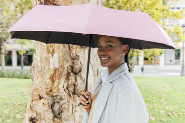 Lächelndes Mädchen mit Regenschirm an einem Baum im Park - JRVF02326