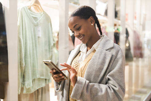 Lächelndes Mädchen mit Smartphone am Schaufenster eines Einkaufszentrums - JRVF02348