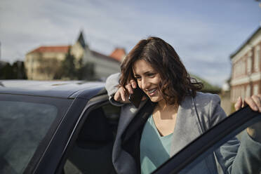 Smiling woman talking on mobile phone at car door - ZEDF04309