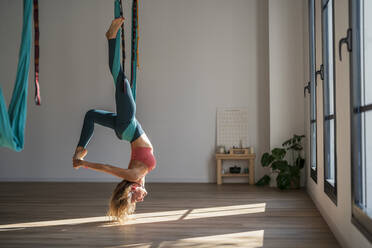 Woman hanging upside down on aerial silk practicing yoga at health club - DLTSF02488
