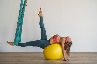 Frau liegt auf einem Fitnessball und dehnt sich mit Luftseide im Yogastudio - DLTSF02491
