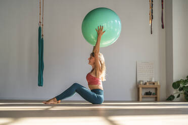 Frau, die im Yogastudio sitzend mit einem Fitnessball trainiert - DLTSF02493