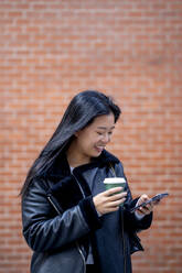 Smiling woman with disposable cup using mobile phone in front of wall - OCMF02299