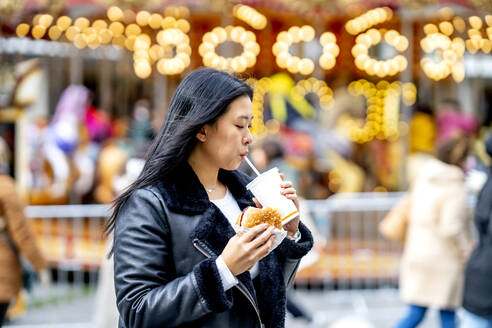 Frau hält einen Burger und trinkt aus einem Einwegbecher im Park - OCMF02307