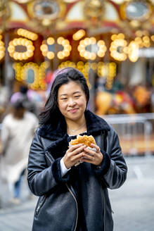 Smiling young woman holding burger at amusement park - OCMF02308