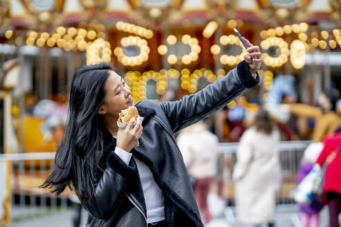 Woman holding burger and taking selfie through smart phone at park - OCMF02310