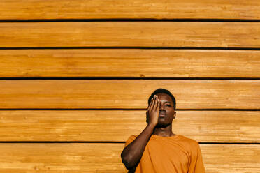 Man with hand covering eye in front of orange wall at sunset - TCEF02133