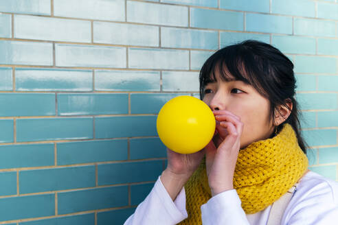 Woman blowing yellow balloon by turquoise brick wall - ASGF02006