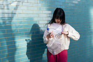 Young woman looking into mesh bag with empty plastic bottles in front of brick wall - ASGF02017