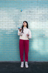 Junge Frau jongliert mit kleinen Kugeln vor einer türkisfarbenen Backsteinwand - ASGF02022